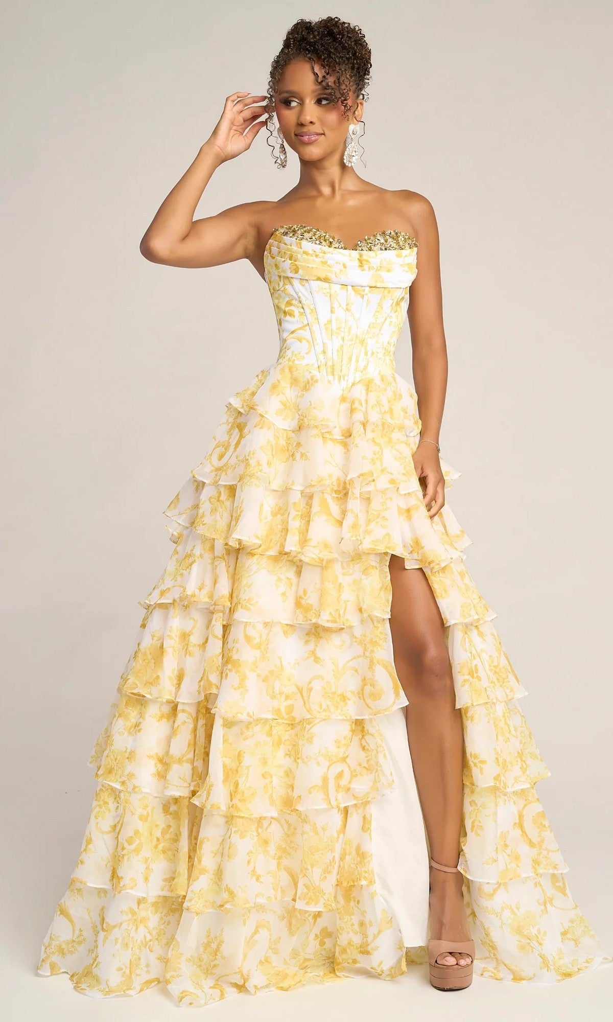 A woman stands against a plain background wearing the Portia and Scarlett PS26210 long prom dress in yellow, a strapless gown with ruffled tiers, floral patterns, and a high slit.